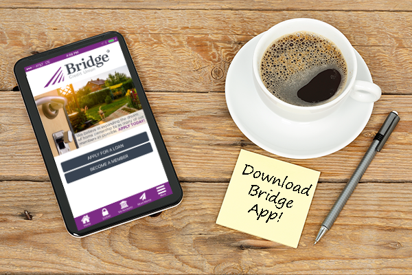 An electronic tablet showing the Bridge app, a cup of coffee on a saucer, a pen, and a paper note pad laying on a wooden background